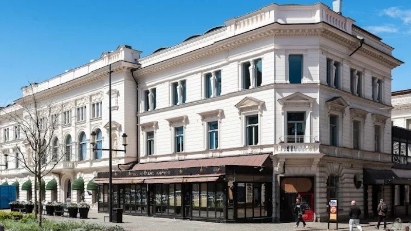 Ståtlig vit stenbyggnad i klassisk arkitektur vid Stora Torget i Trollhättan, med restaurang i bottenplan och blå himmel som bakgrund.